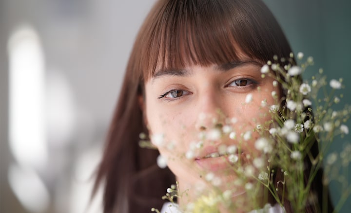 Una fotografía de retrato de una mujer con flequillo y cabello suelto, la cual mira directamente a la cámara, con un hermoso efecto bokeh creado por la presencia de flores de gypsophila delante de su rostro.